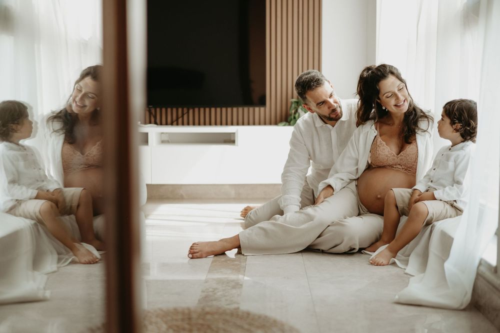 Pregnant woman sitting on floor with partner and toddler in bright modern room during maternity photo session.