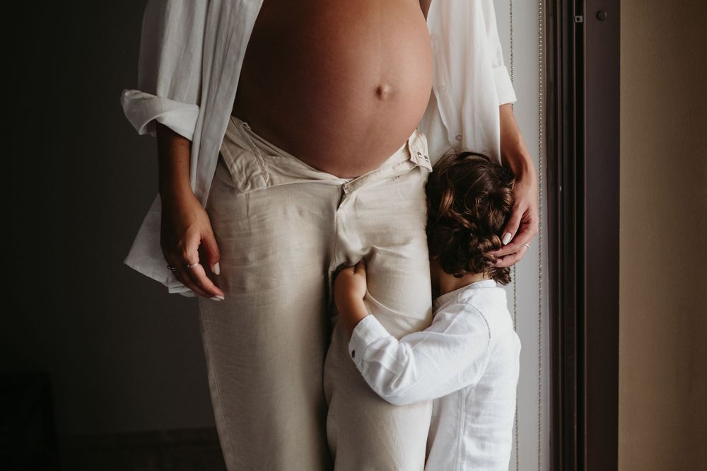 A young child hugging a pregnant mother's belly near a window, both dressed in white linen clothing.