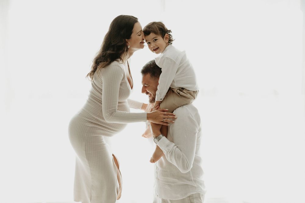 Pregnant woman in white dress with partner and toddler on shoulders sharing a joyful family moment against white background.