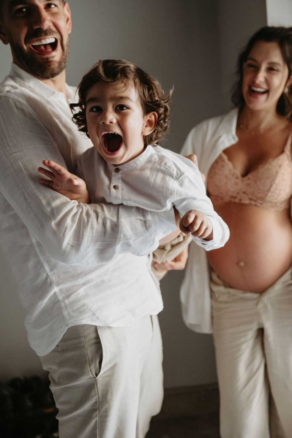 Joyful family maternity photo with father holding excited toddler in white outfit, pregnant mother smiling in background.