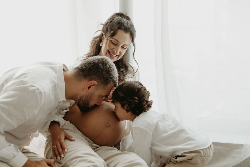 Happy family with parents kissing their toddler child on a bright white bed in a warm, intimate moment.