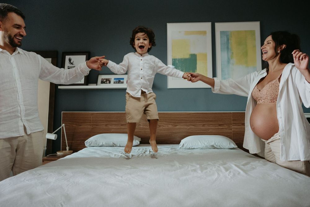 Happy family with pregnant mother and father holding hands with excited child jumping on bed at home.
