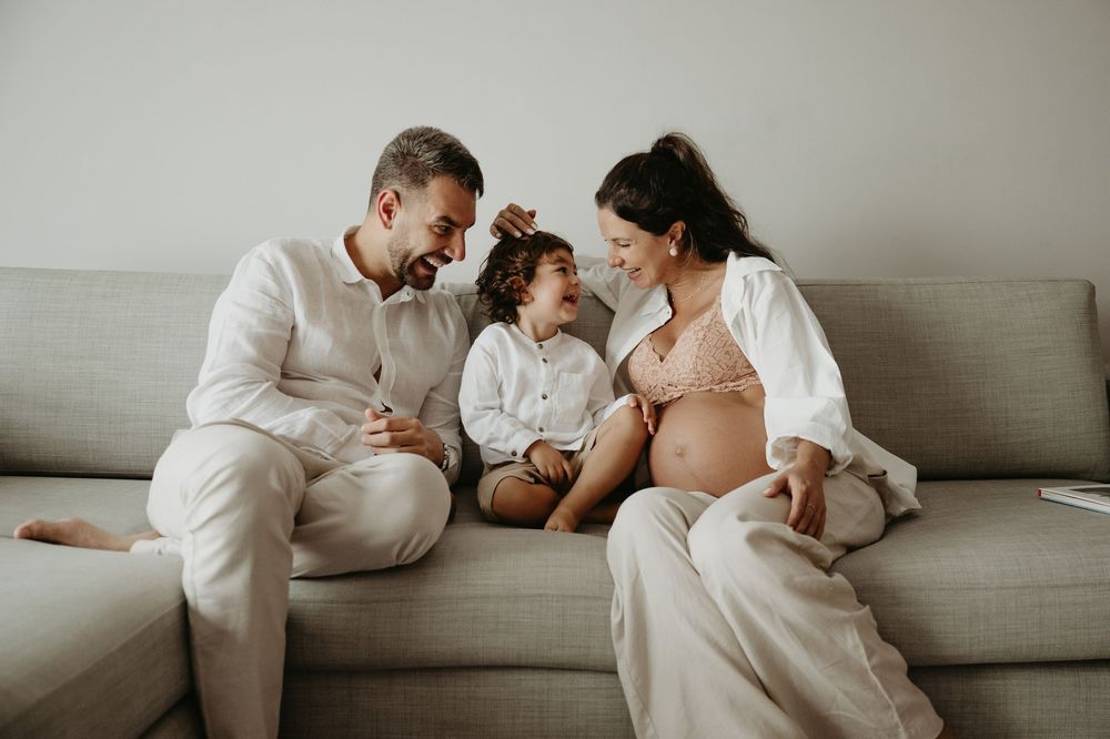 Pregnant woman sitting on sofa with husband and toddler child, happy family portrait at home.