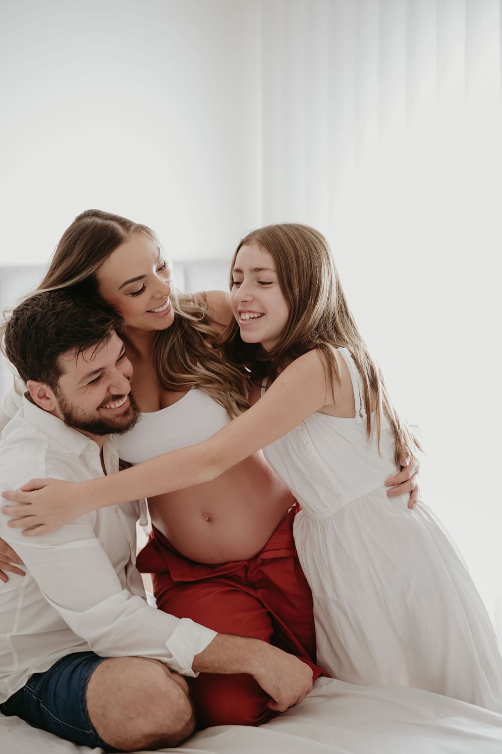 Pregnant woman in red pants sits on bed with husband and daughter sharing a joyful family embrace.
