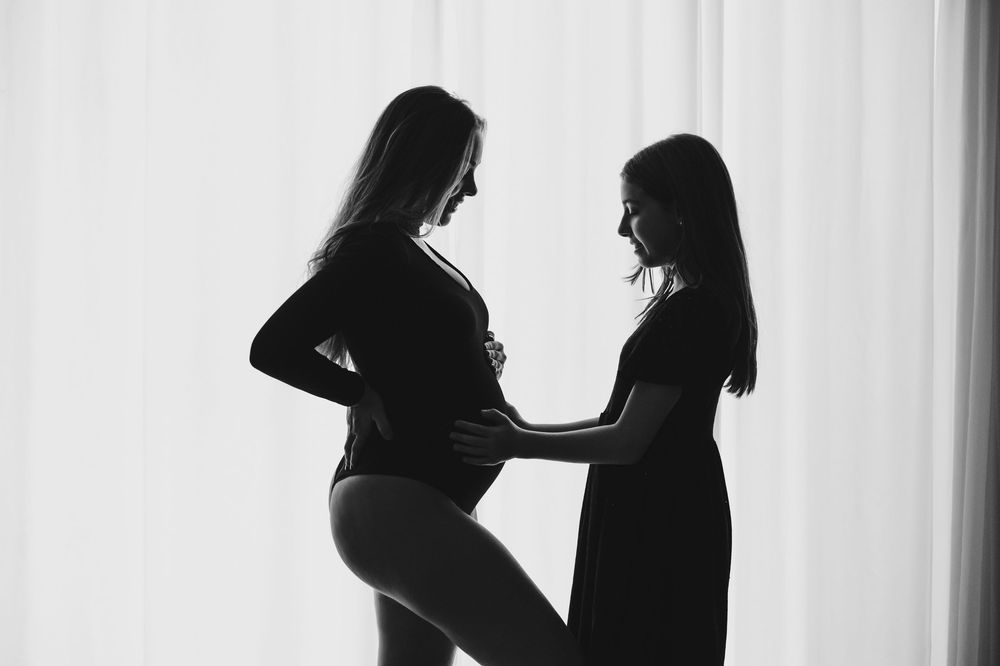 Black and white silhouette of pregnant woman and friend touching baby bump near sheer curtains.