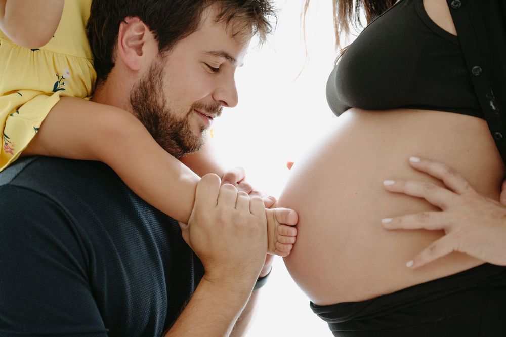 A bearded man tenderly holds a child's foot near a pregnant woman's bare belly in a warm family maternity photo.