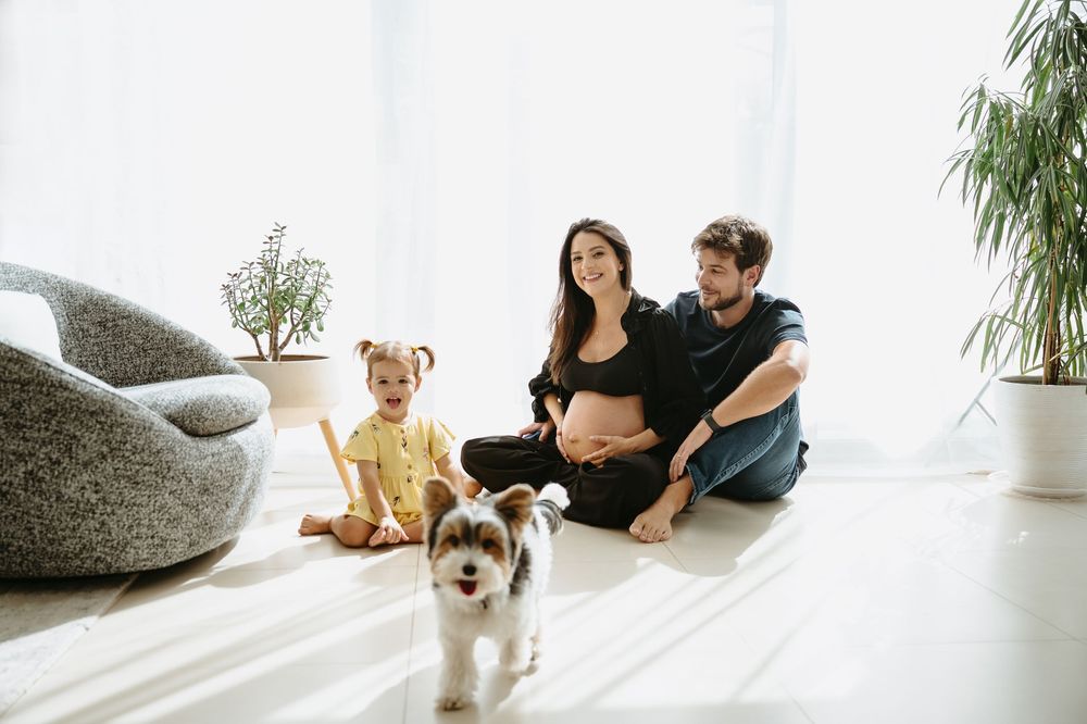 Pregnant woman sits on floor with husband, toddler daughter, and playful puppy in bright white living room.