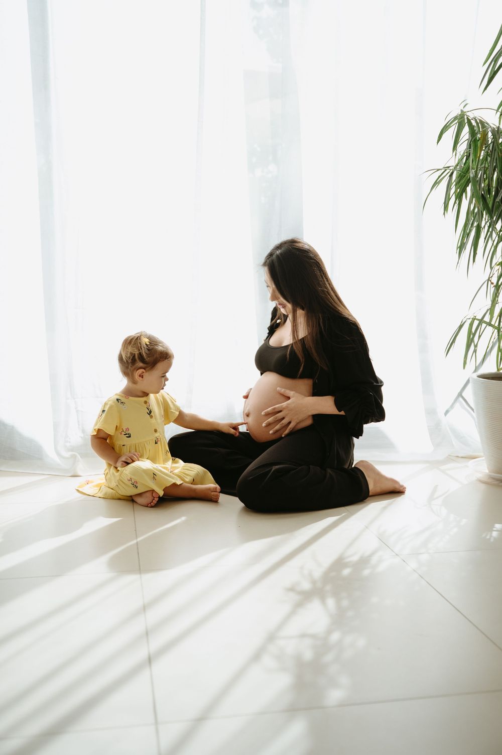 Pregnant woman in black outfit sitting on floor with toddler in yellow dress, sharing a tender moment indoors.