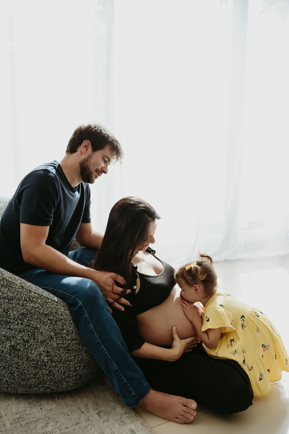 A pregnant woman sits with her husband and young daughter who kisses her baby bump in a bright, cozy room.