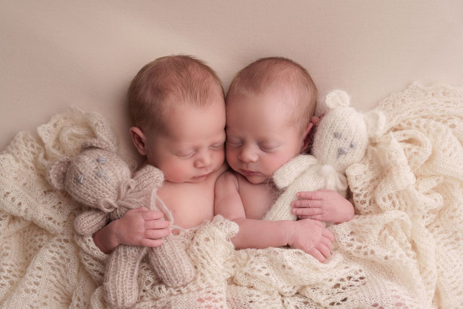 Two sleeping newborns cuddle together on cream knitted blankets in a tender studio portrait.