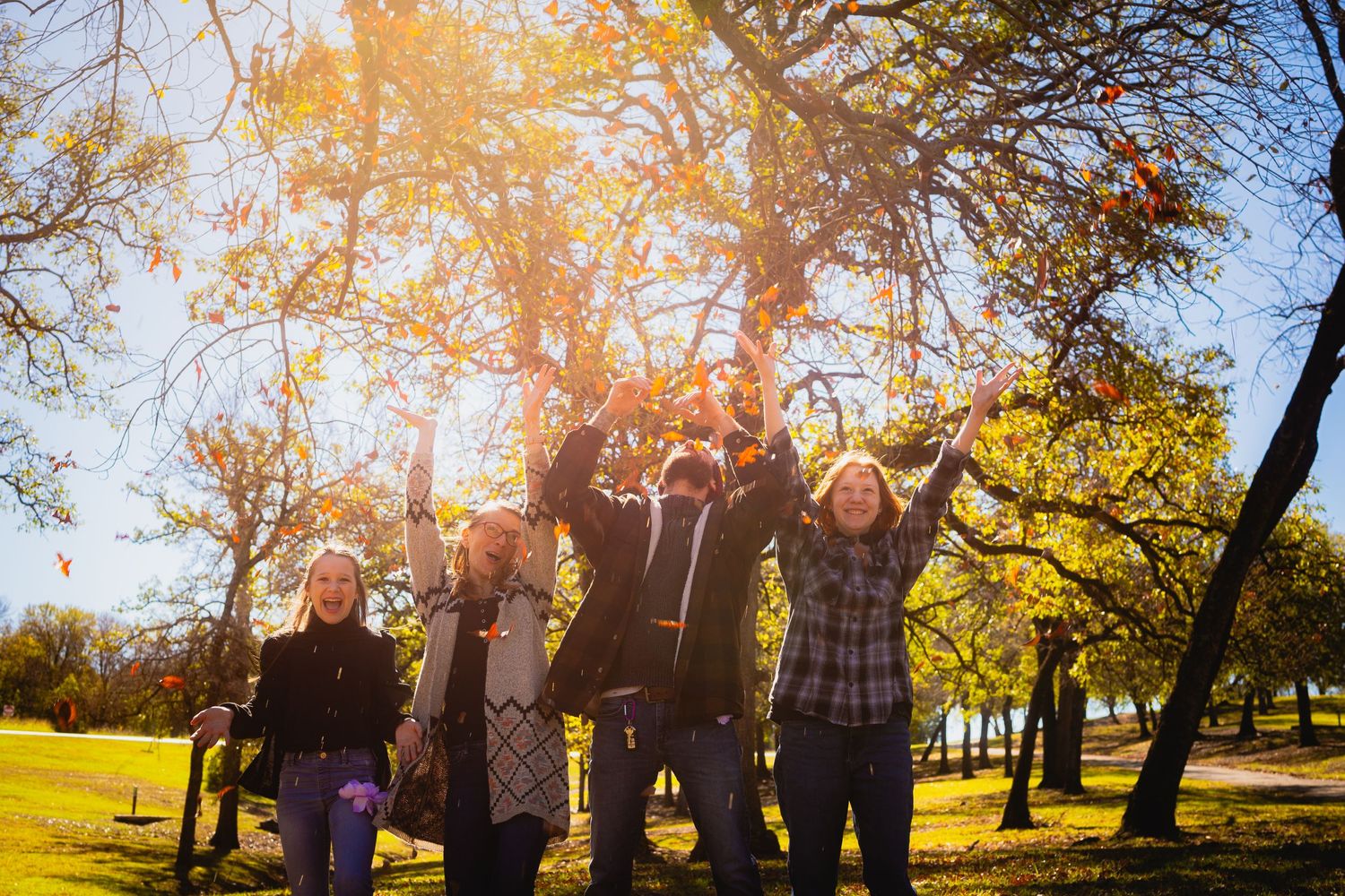 Group celebrating with raised arms in a sunny park with autumn trees.