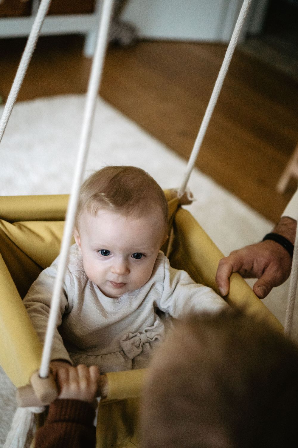 Baby in a yellow indoor swing wearing a light colored sweater, viewed through swing ropes.