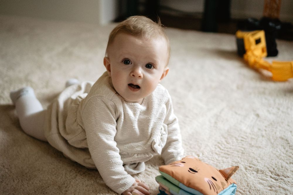 Small infant in cream colored sweater lying on carpet with a toy book and yellow toy in background.