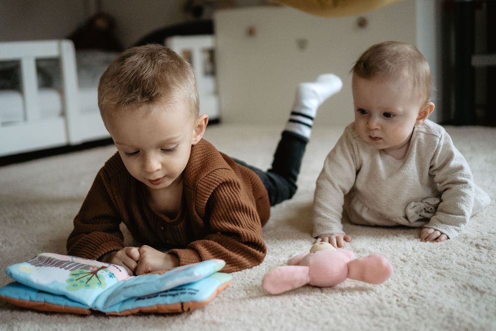 Two young children lying on a white carpet at home while looking at a children's book.