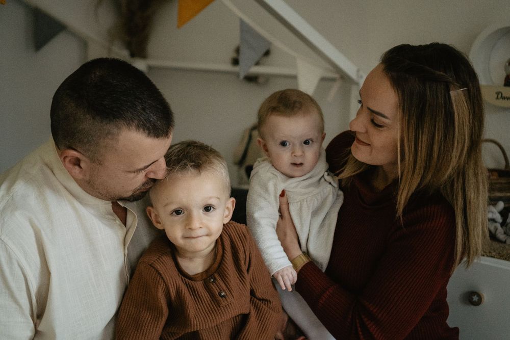 A cozy indoor family portrait shows parents and two young children together in warm autumn-toned clothing.