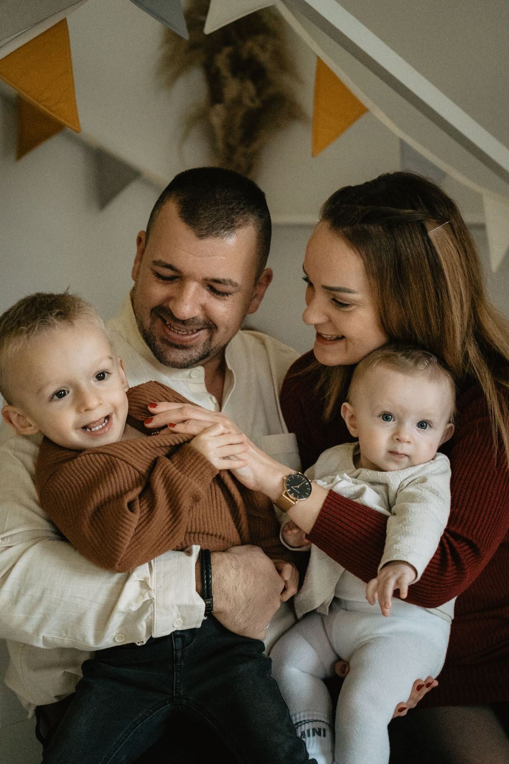 Cozy indoor family portrait showing parents cuddling with two young children under festive yellow triangle bunting decorations.