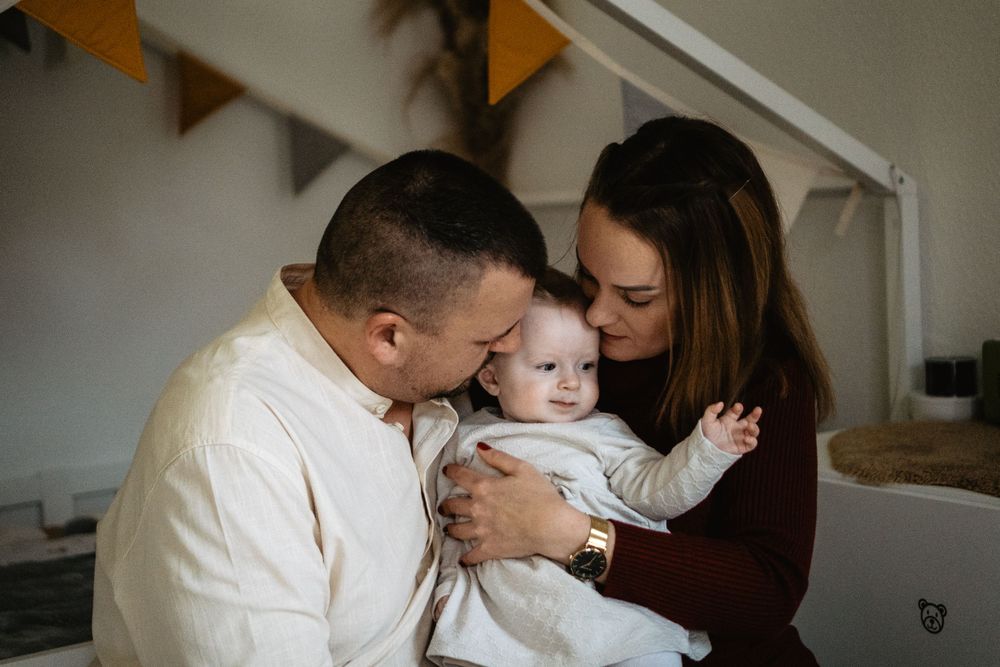 A loving couple sharing a tender moment with their baby in a cozy home setting with bunting decor visible in background.