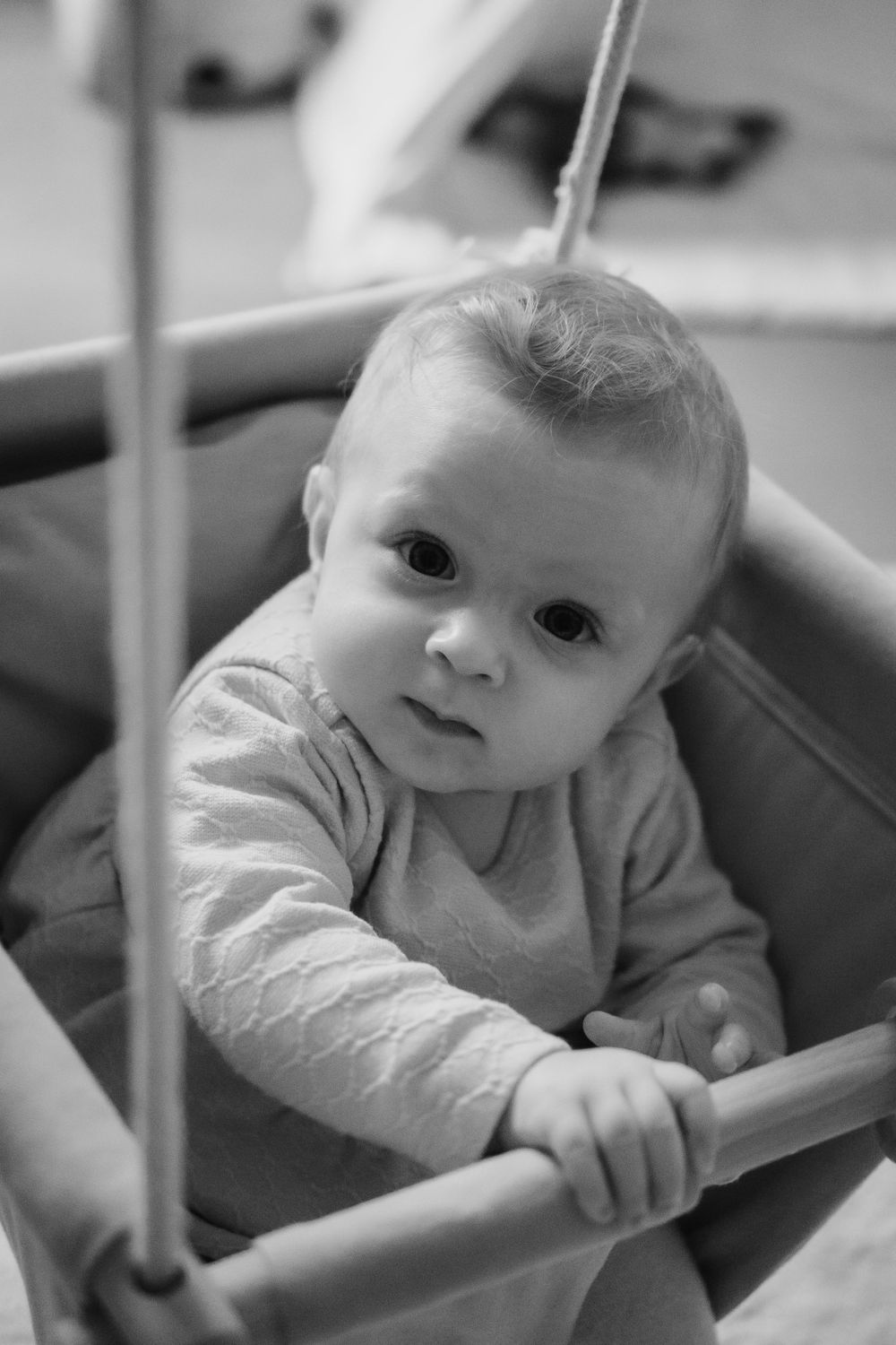 A baby in a light colored sweater leans against what appears to be part of a crib or play structure in black and white.