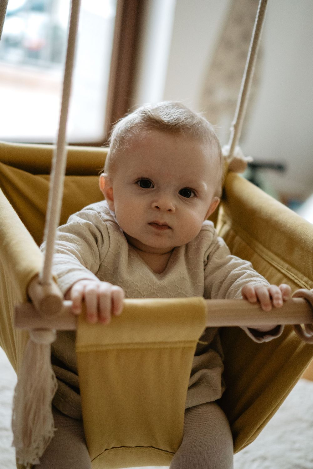 Baby sits in a tan fabric hammock swing while holding onto the edge in a cozy indoor setting.