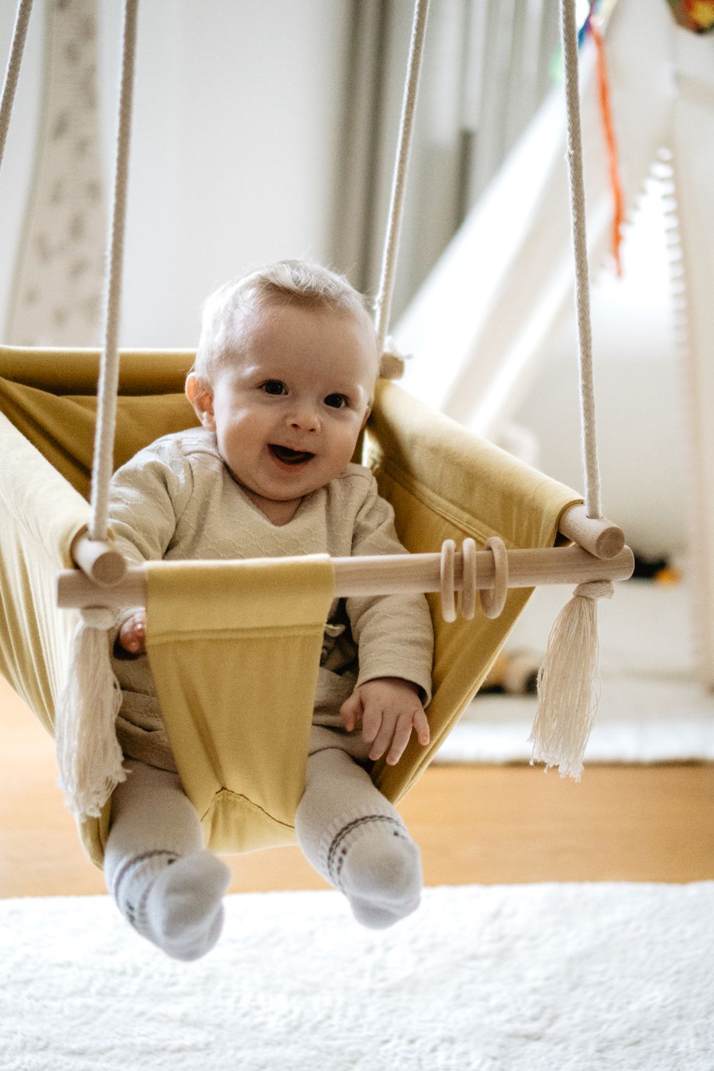 Baby enjoys playtime in a cozy beige fabric swing indoors near a bright window.