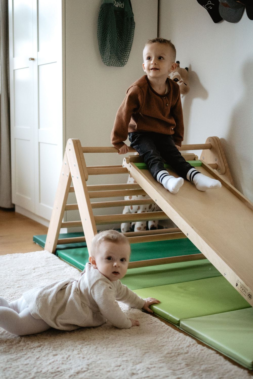 Two toddlers play on a wooden climbing structure with ramp and mats in a minimalist playroom setting.