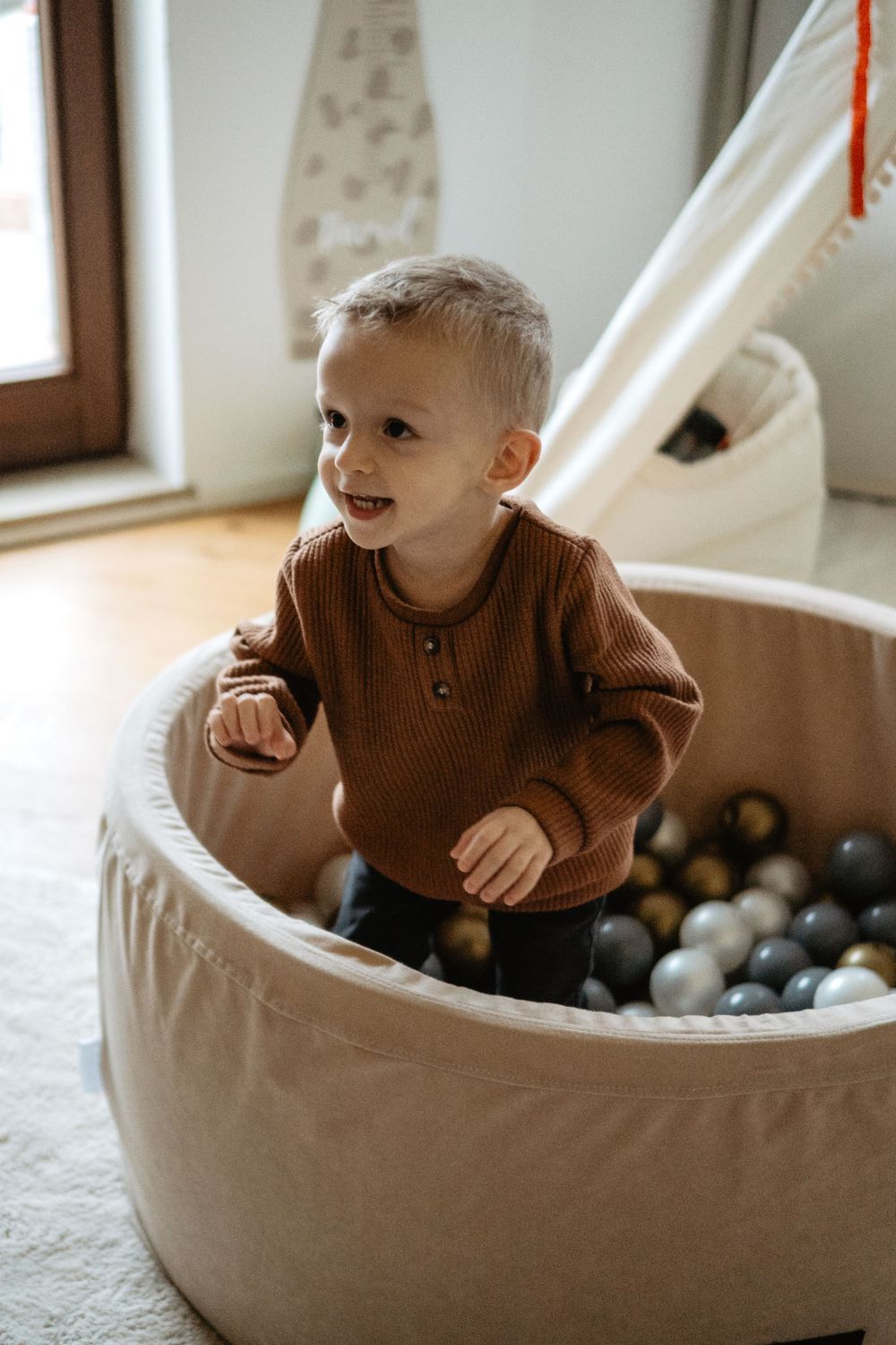 Child in brown sweater playing in a cream-colored ball pit filled with neutral-toned balls in a cozy home setting.
