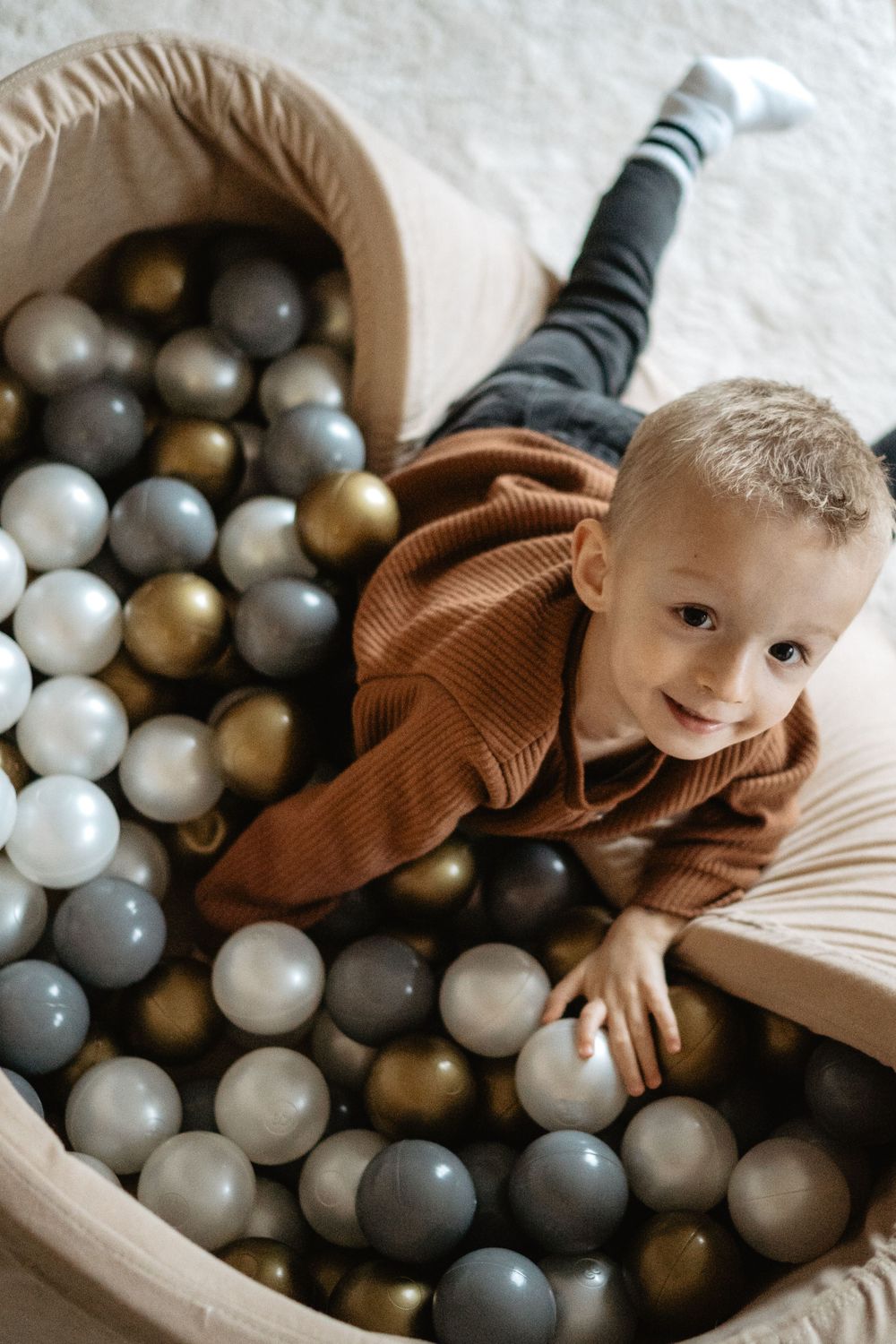 Child playing in a round ball pit filled with metallic grey, white and gold plastic balls while wearing a brown sweater.