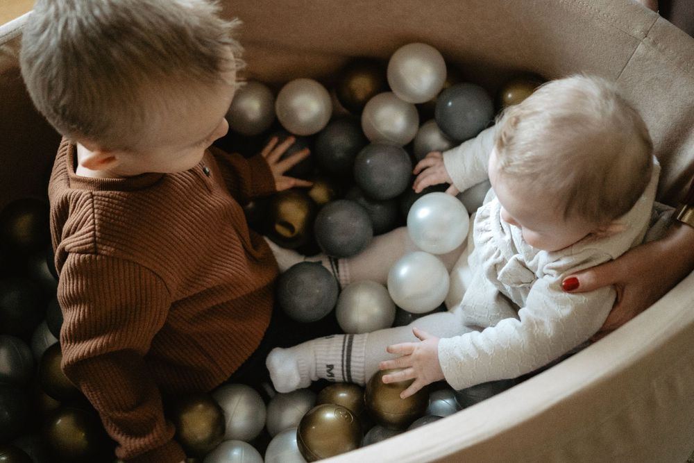 Two small children play in a neutral-toned ball pit filled with gold, white, and gray spheres.