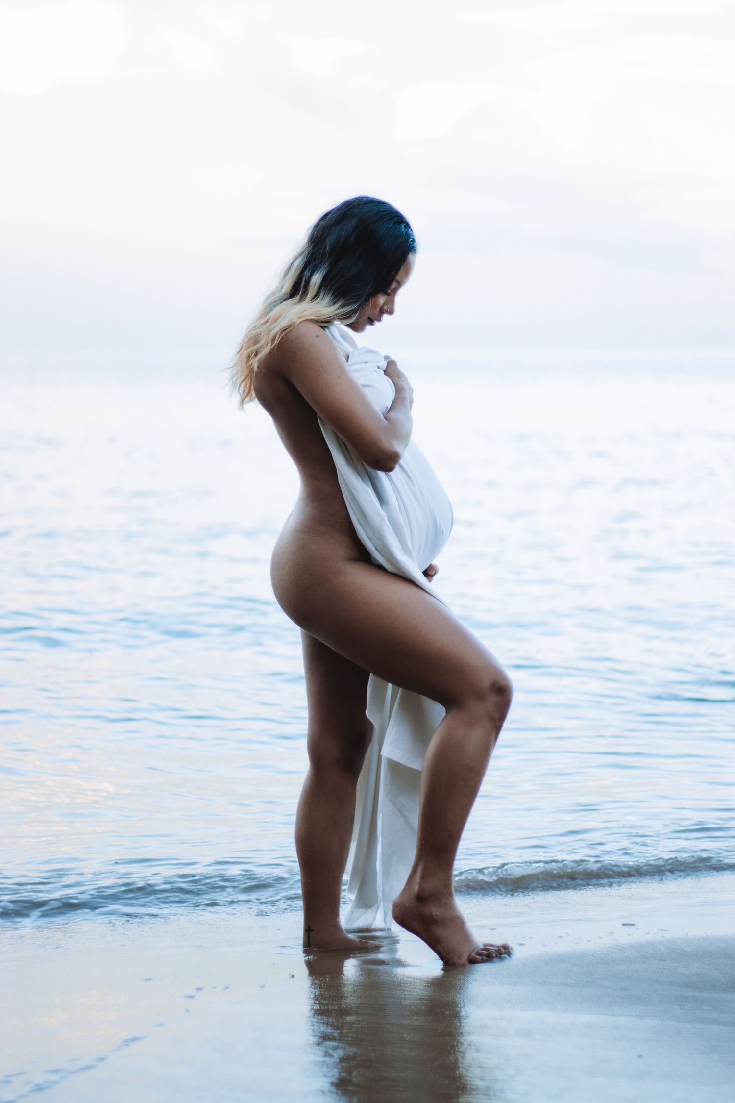 Model centre of frame, dark skin, Balayage hair black to blonde, pregnant using a white sheet to cover breasts and belly
