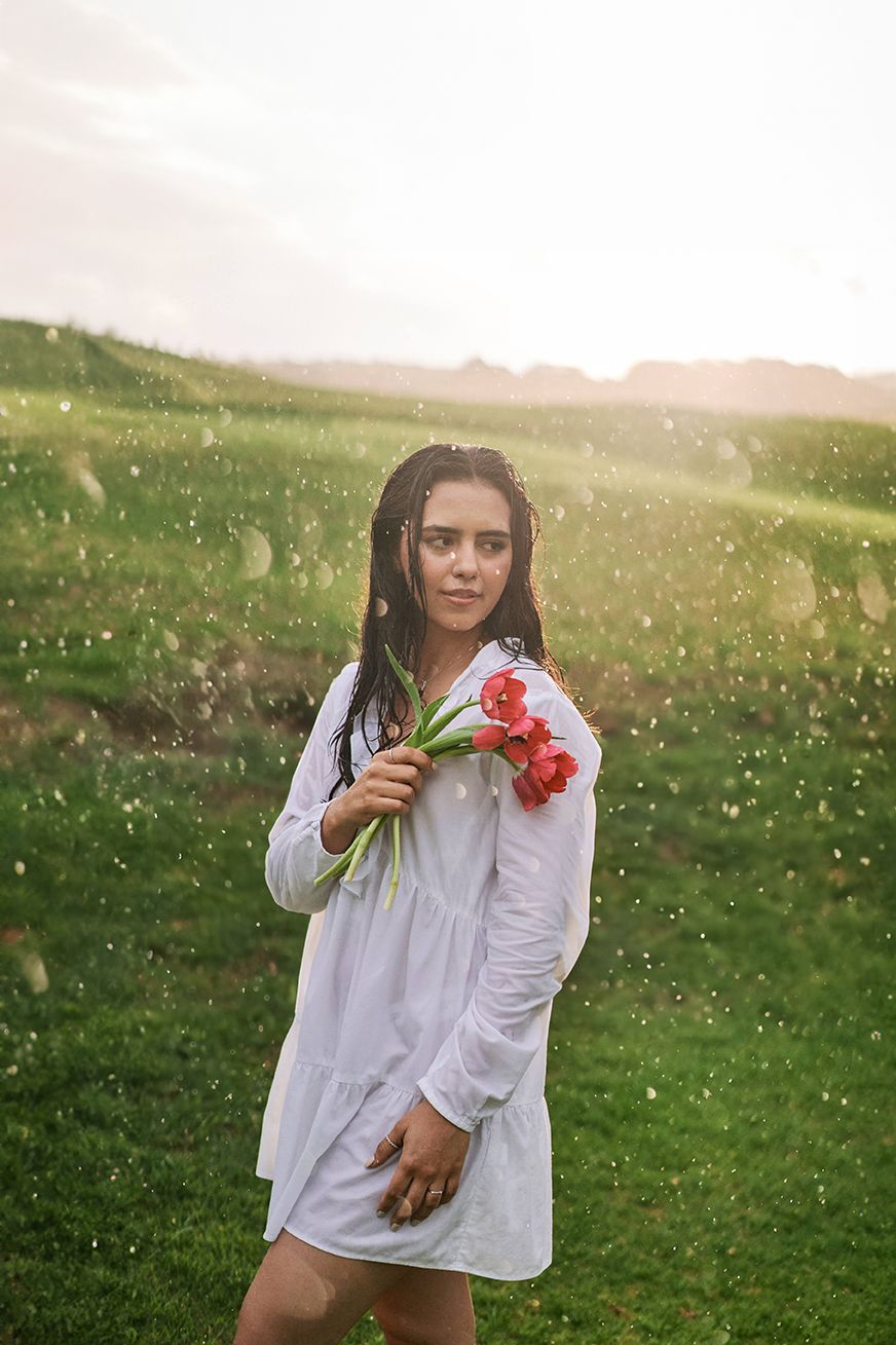 Woman in white dress holding red flowers in sunlit meadow with bokeh effect.