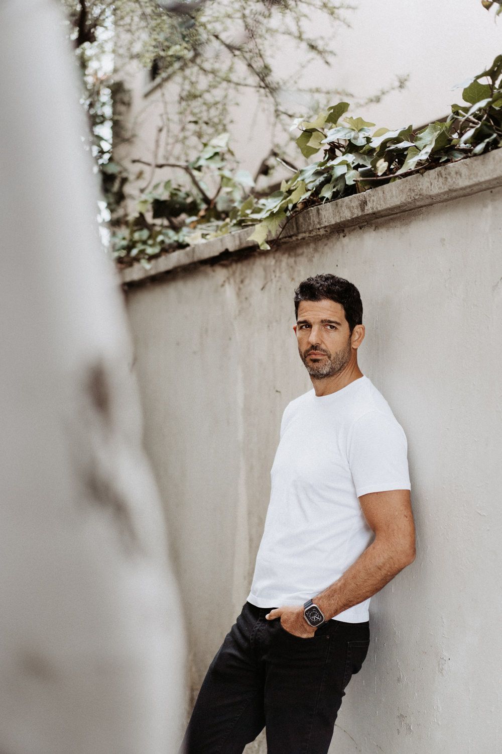 A person in white t-shirt and dark pants poses against a textured wall with climbing vines.