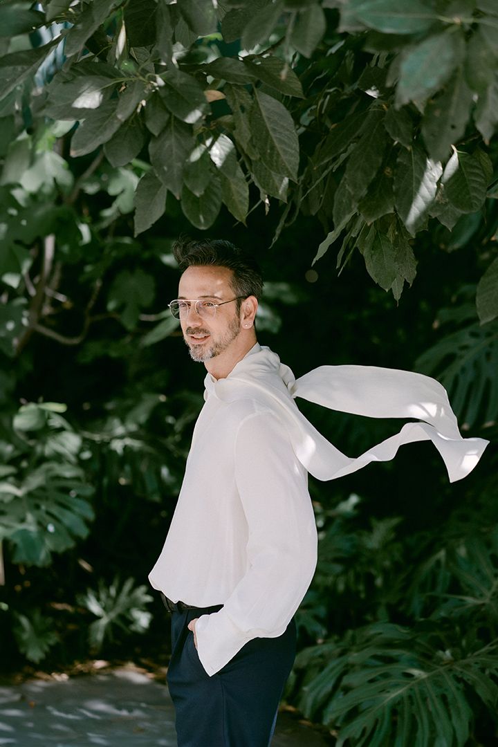 A person in a flowing white shirt poses against tropical foliage with fabric billowing in the wind.