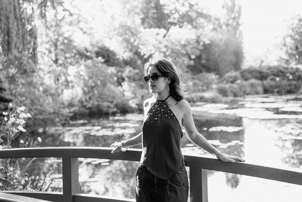 A person in sunglasses stands on a wooden bridge over a serene pond in black and white.