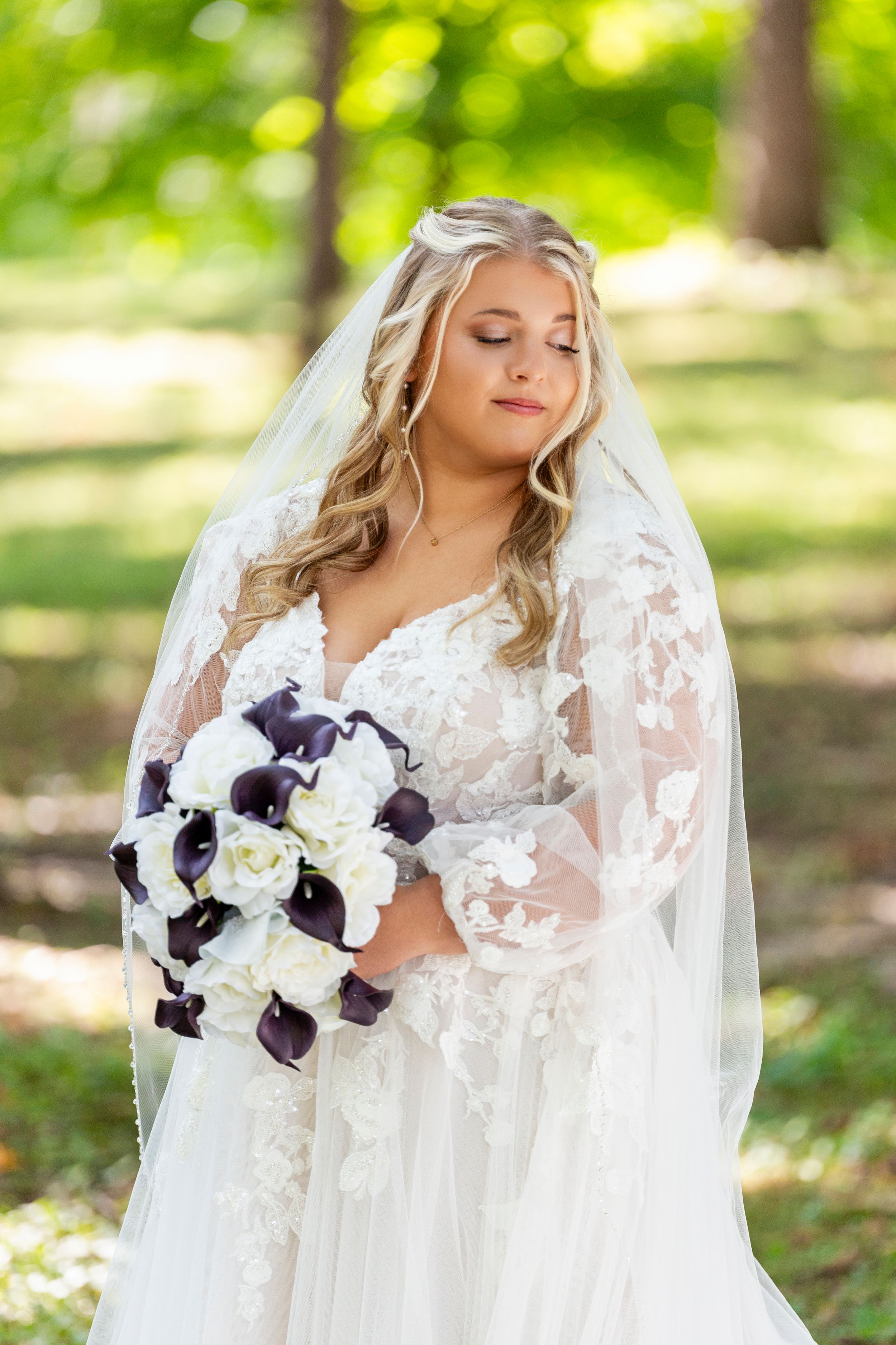 Bride in lace gown and veil holds purple and white bouquet in sunlit garden setting.