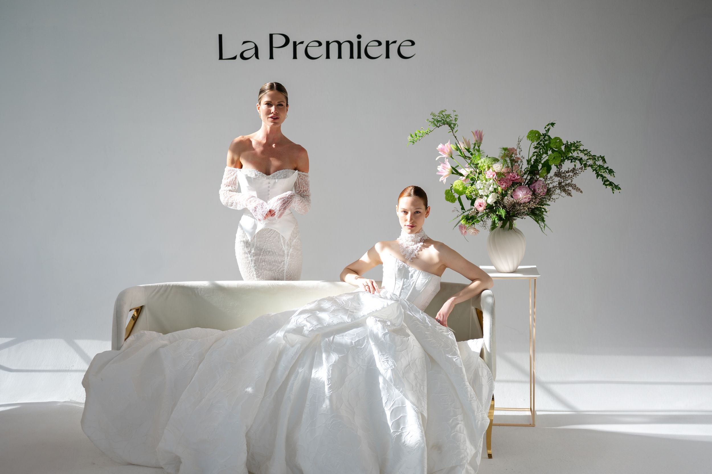 Two elegant white wedding gowns displayed in a minimalist studio setting with greenery decor.