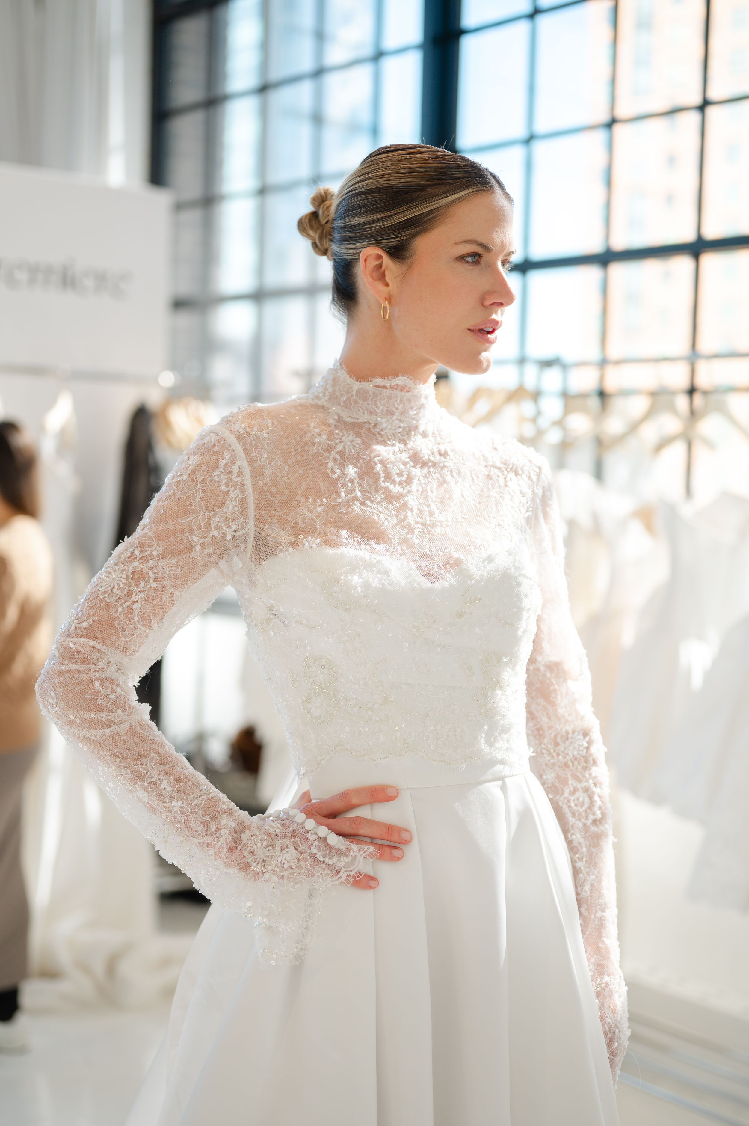 Bride wearing an elegant high-neck long sleeve lace wedding gown with a fitted bodice in a sunlit bridal boutique.