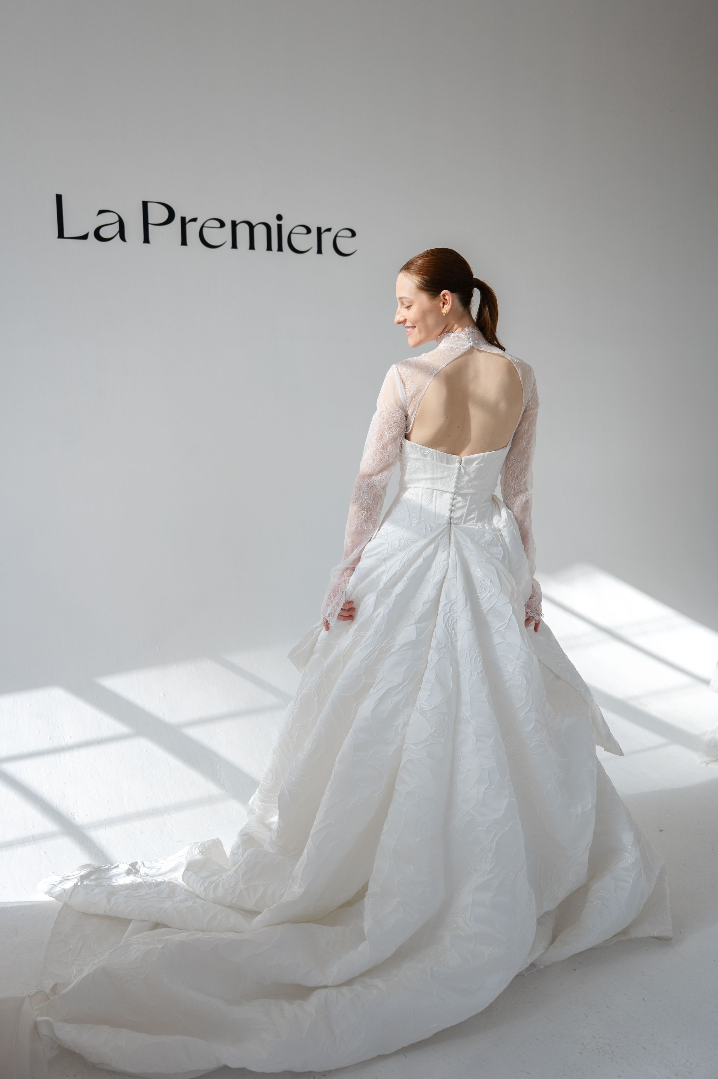 A bride in a backless wedding gown with sheer sleeves poses against a white wall with dramatic shadows.