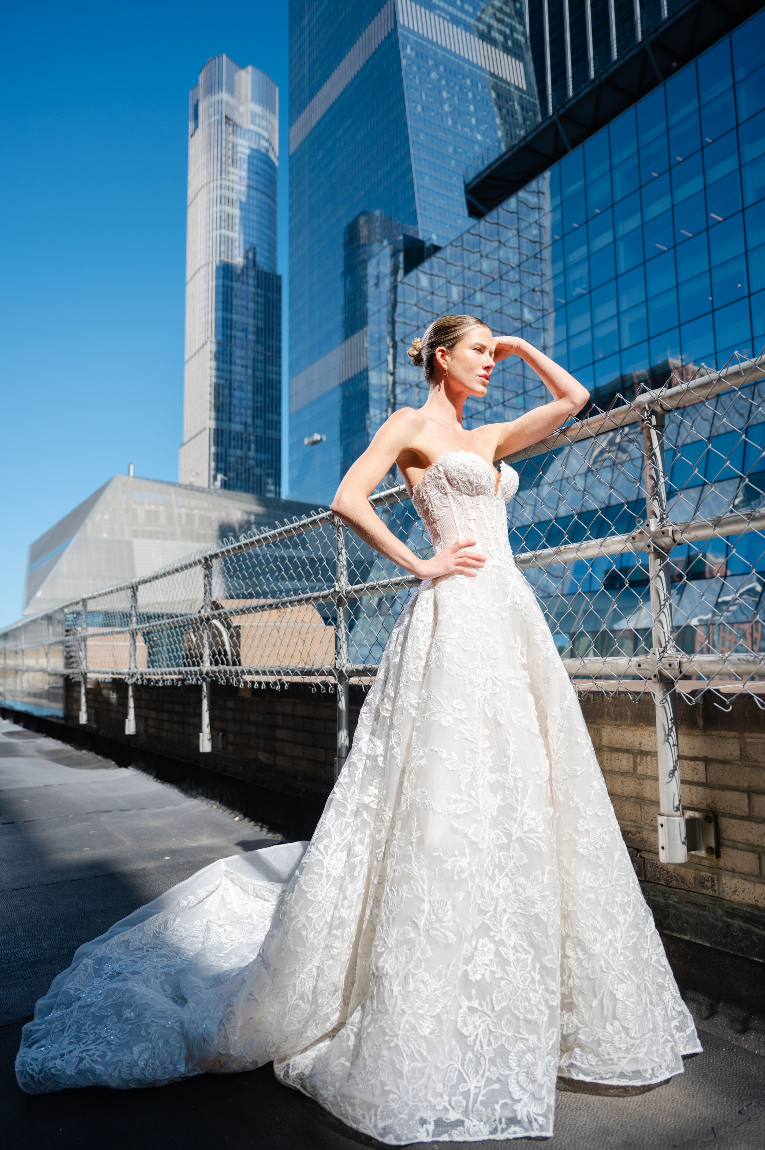 A bride in a white lace ballgown poses gracefully against modern glass skyscrapers in an urban setting.
