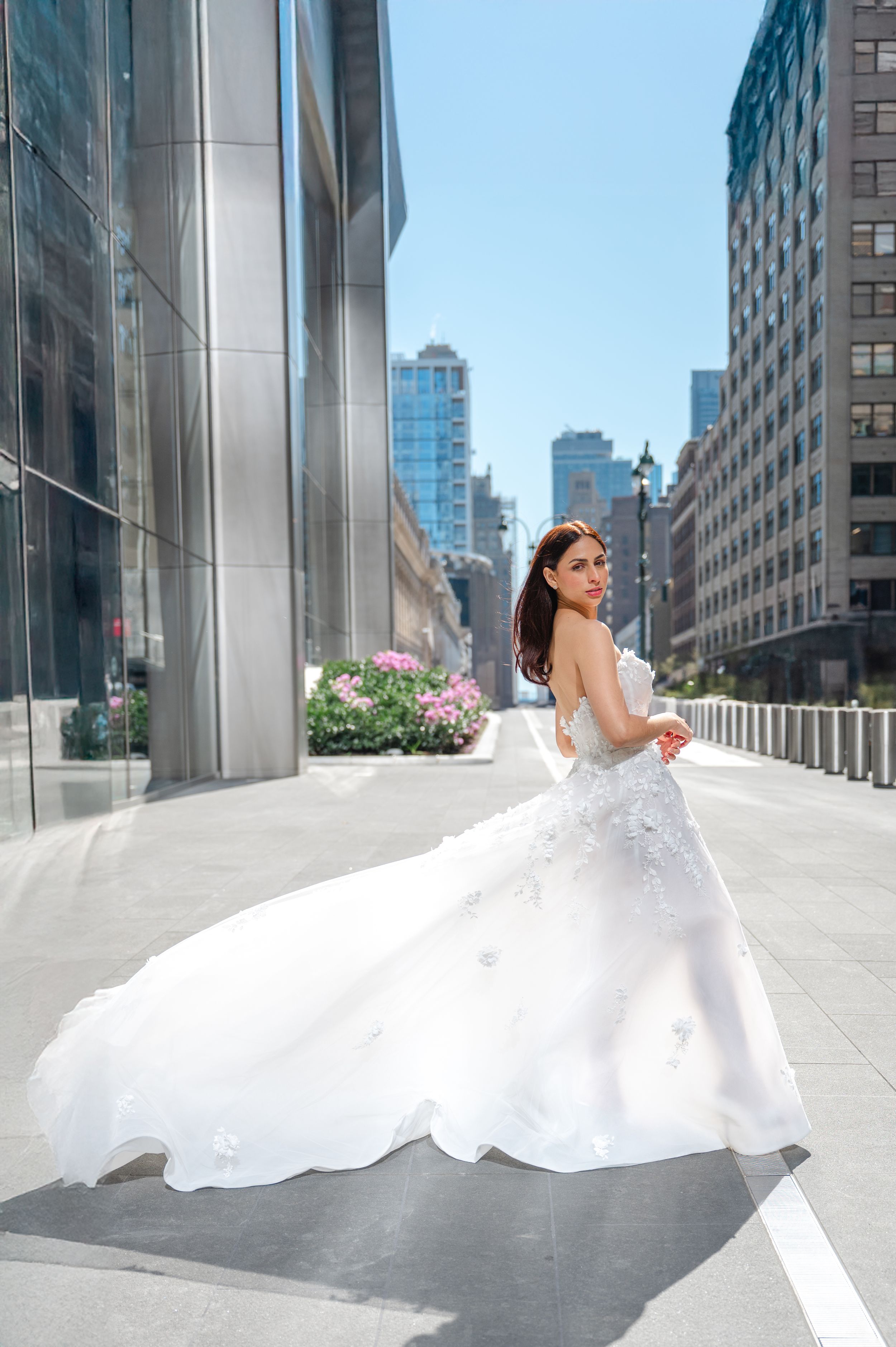 A bride in a flowing white wedding gown twirls on a modern city street among tall buildings and pink flowers.