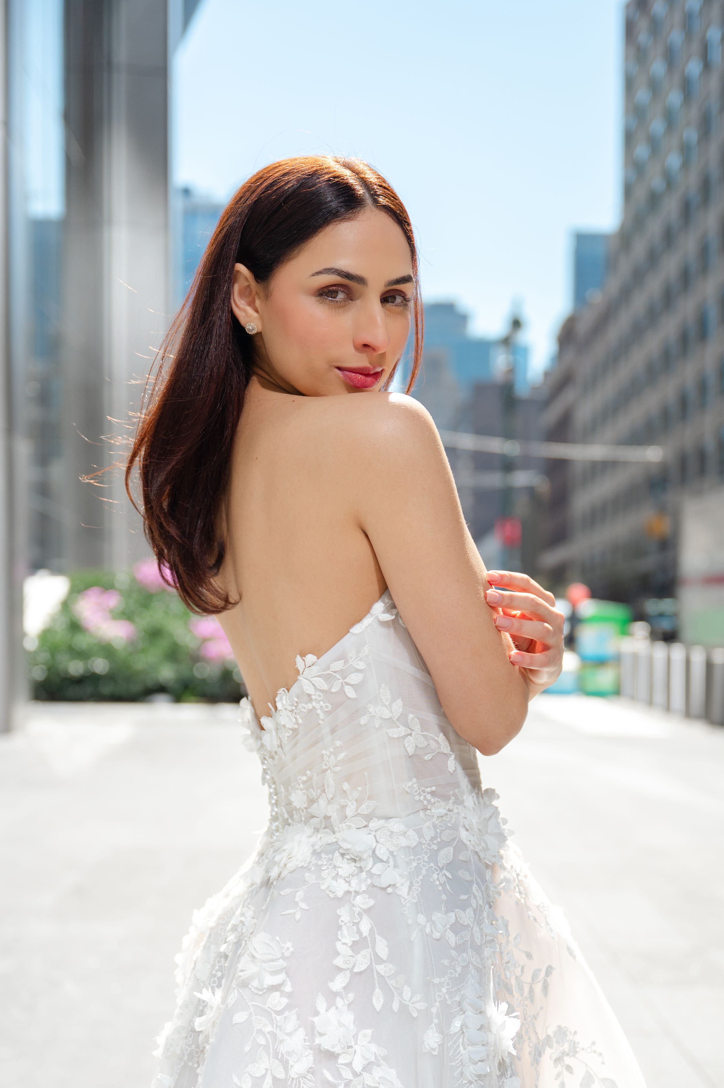 Elegant bride in white floral lace wedding dress poses on city street with skyscrapers in background.