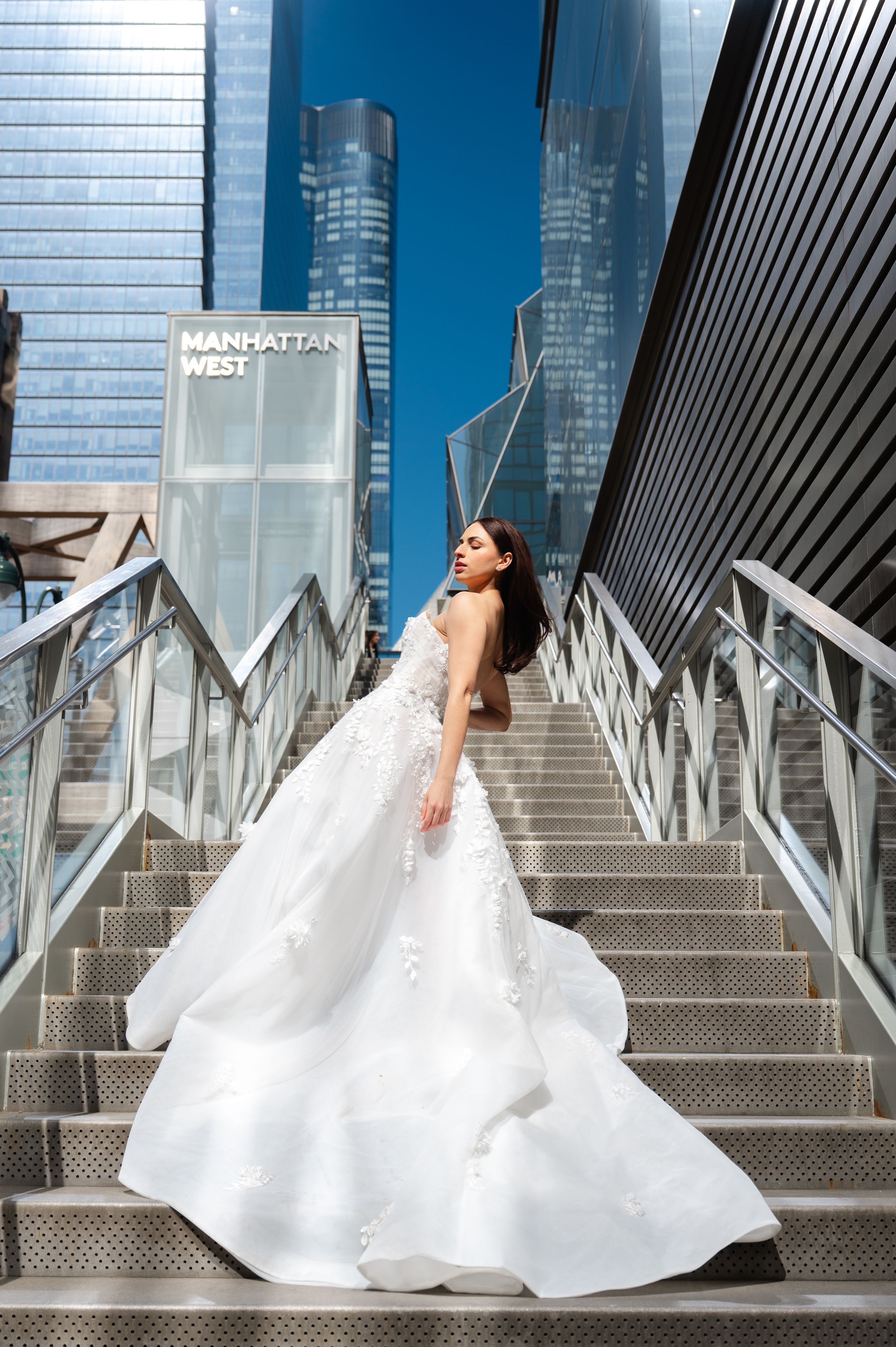 A bride in a flowing white ball gown poses gracefully on modern metal stairs with glass skyscrapers in the background.