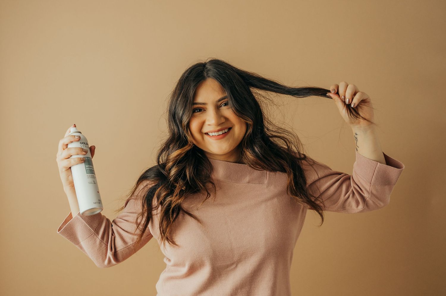 Woman with long hair playfully styles hair with product against beige background.