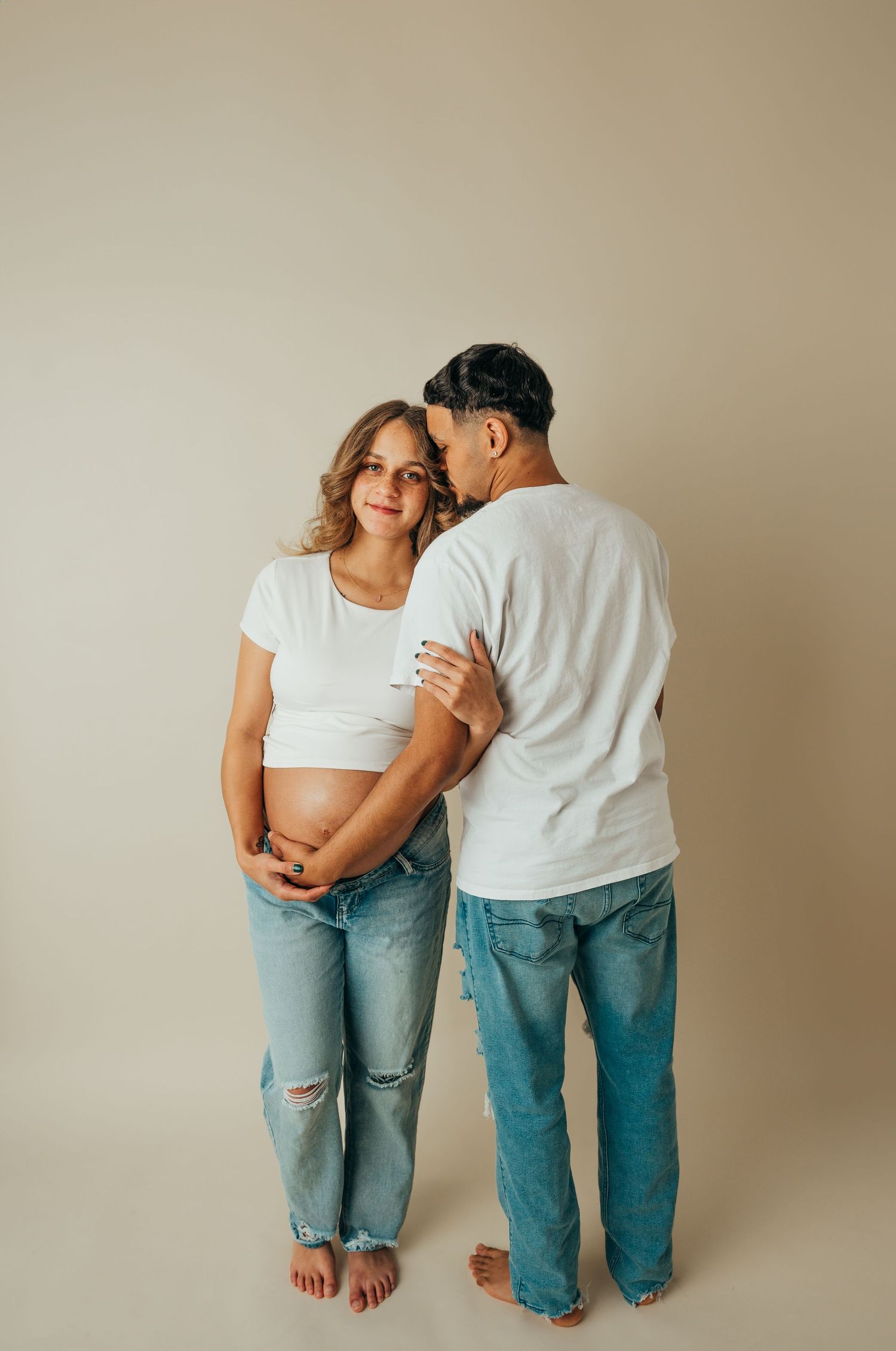 Couple in casual denim and white shirts share intimate moment against neutral background.