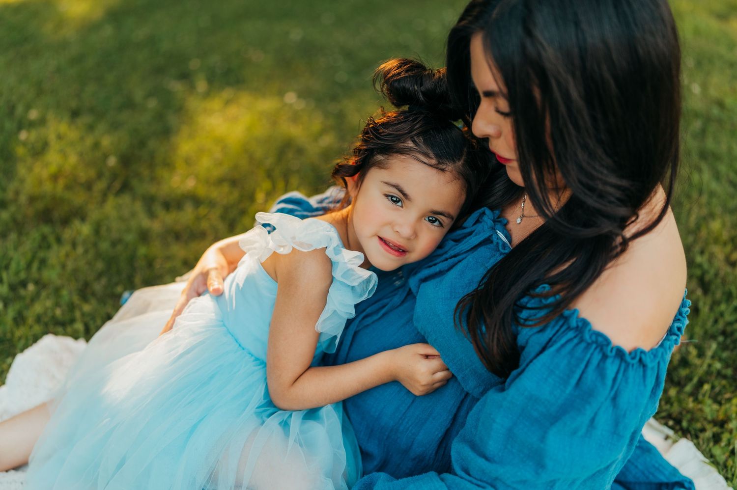 Mother and daughter in matching blue dresses share tender moment on grass.
