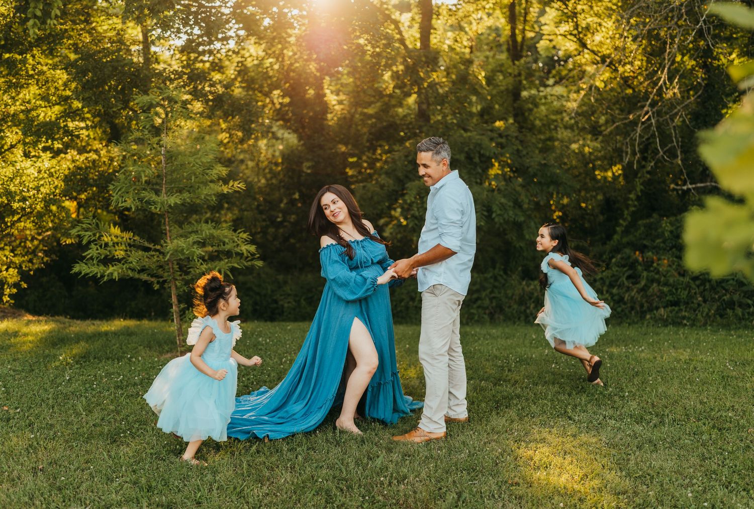 A joyful outdoor maternity photoshoot with family members dancing and twirling in blue dresses at sunset.