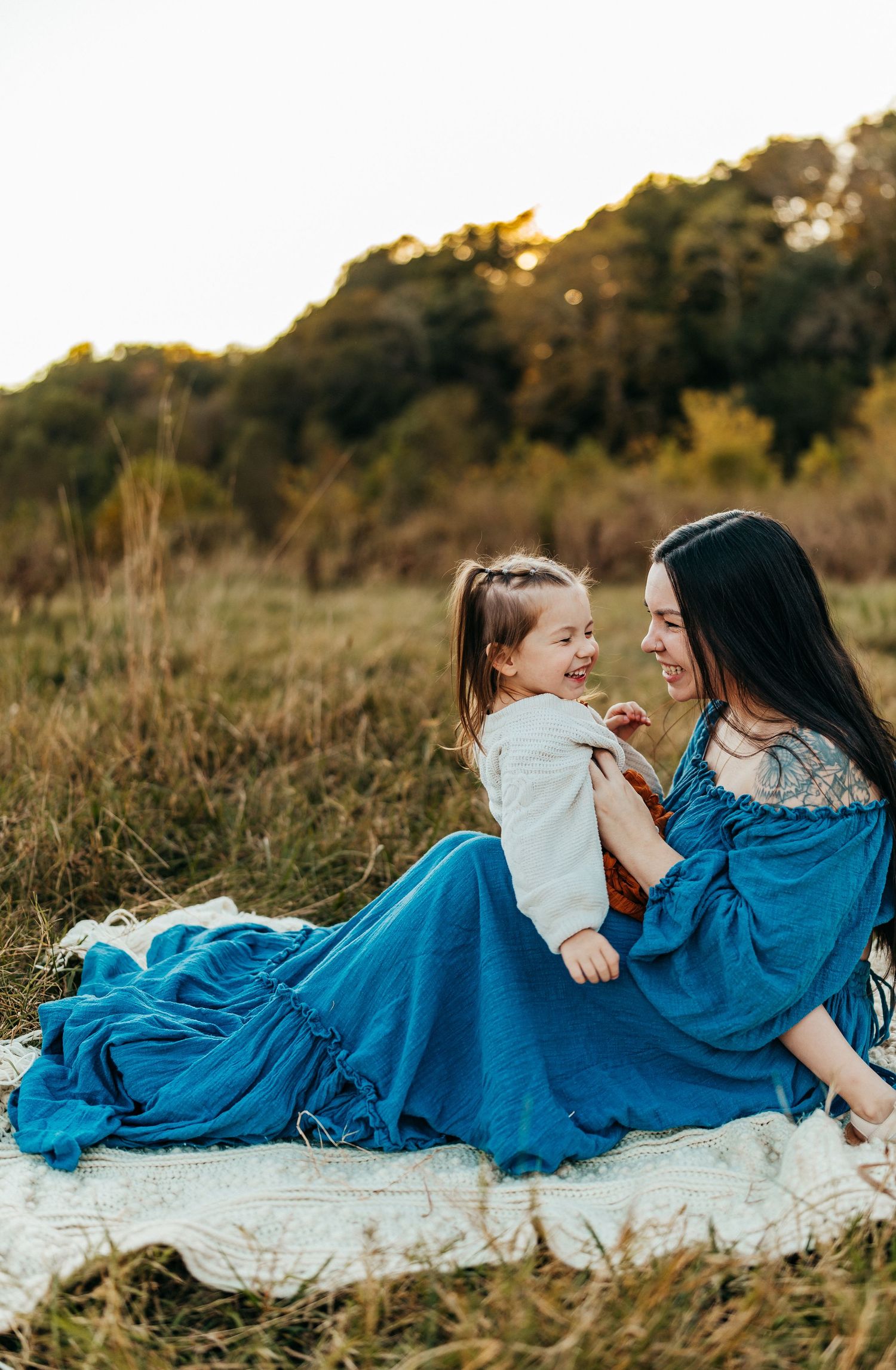A tender maternal moment captured on a blanket in a field at sunset with flowing blue dress. rockford il