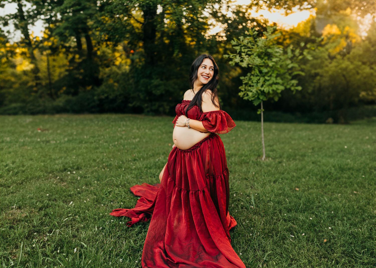 A maternity portrait featuring a flowing red dress in a sunlit green field.