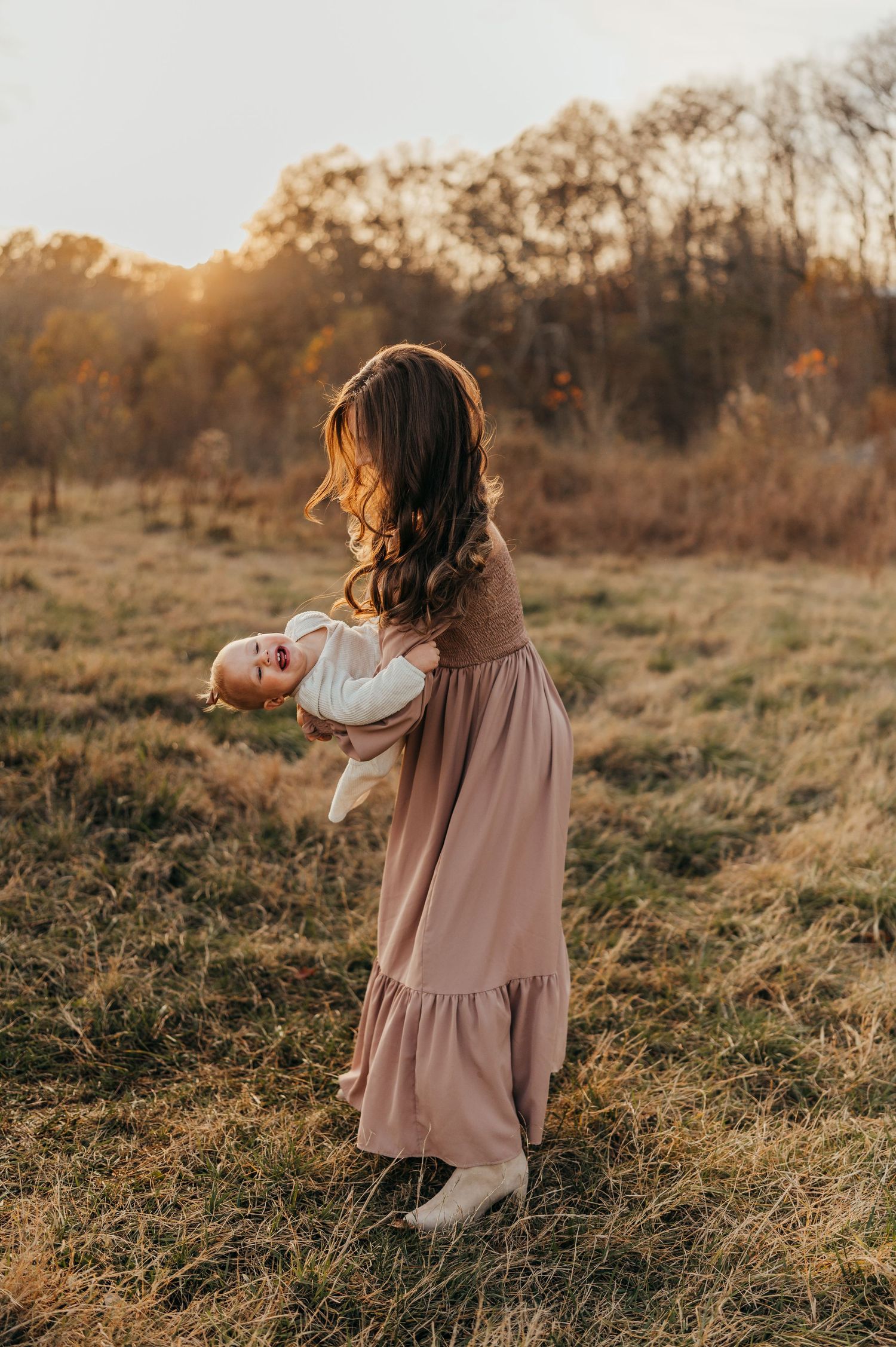 Woman in mauve dress dancing with baby during sunset in rustic field setting.