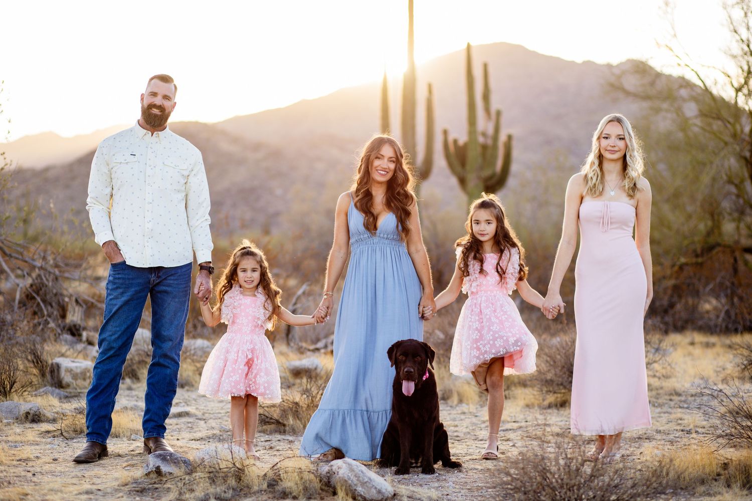 Family with dog poses in desert landscape with saguaro cactus at sunset in Arizona.