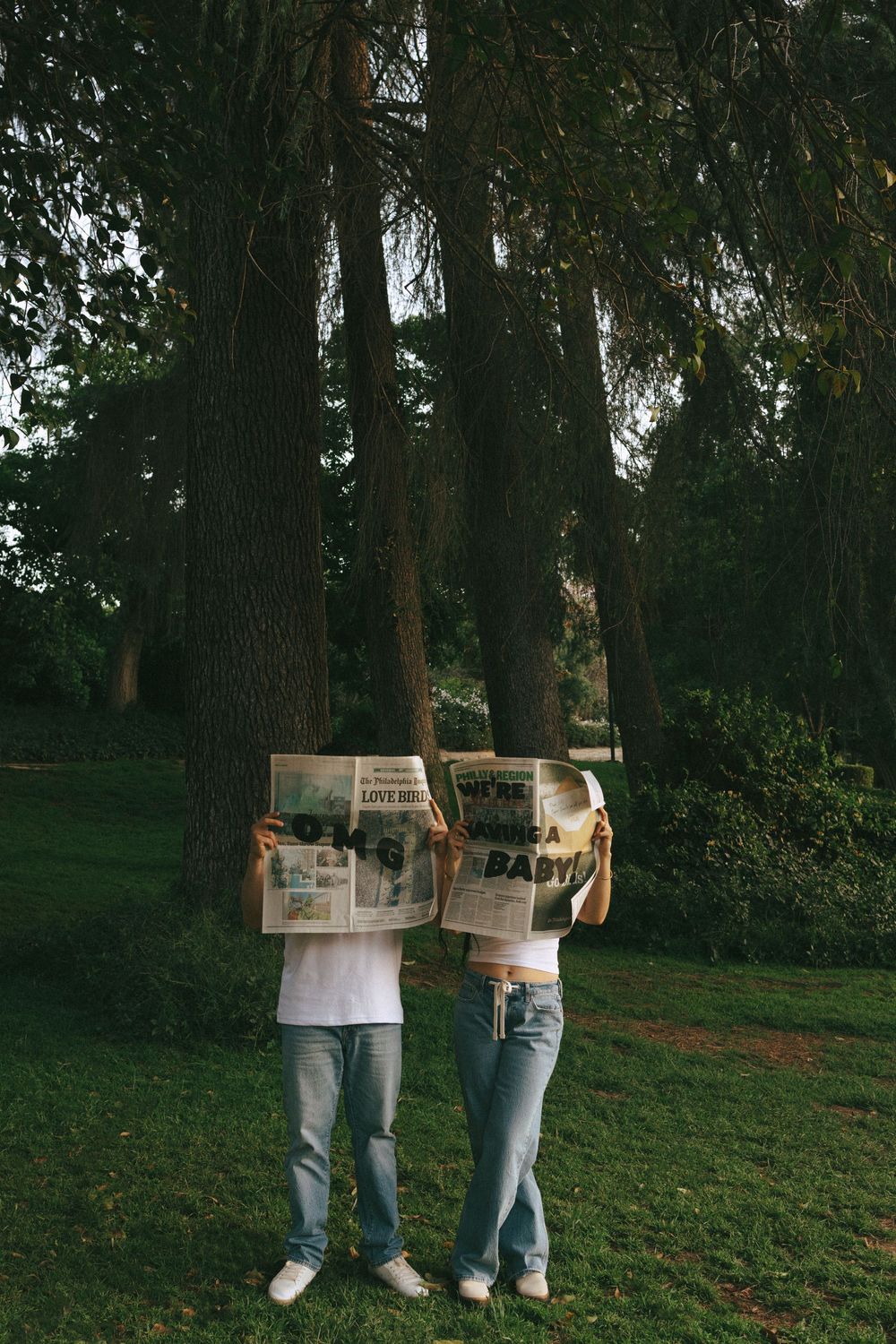 A couple standing in a wooded park holding newspapers up to obscure their faces while wearing casual clothing.